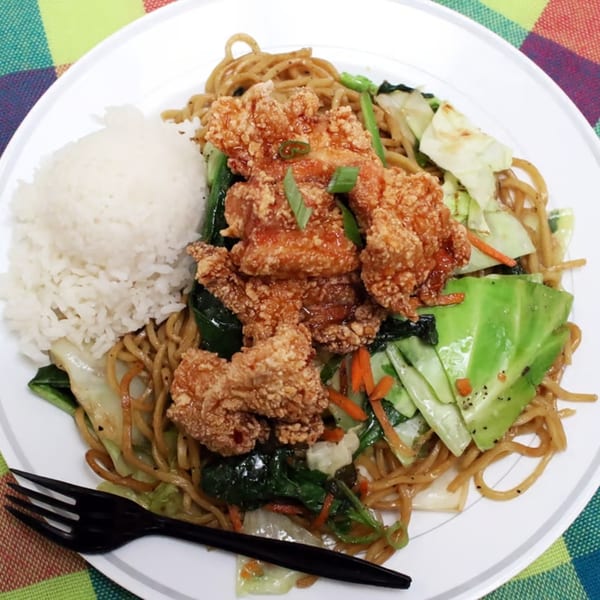 Hawaiian plate lunch with crispy fried chicken, noodles, vegetables, and white rice on a colorful plaid tablecloth.