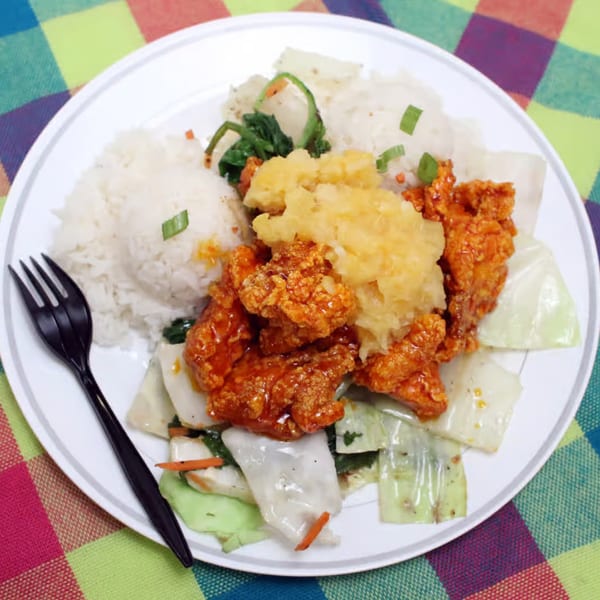 Hawaiian plate lunch with crispy orange chicken, rice, cabbage, and pineapple on a colorful plaid tablecloth.
